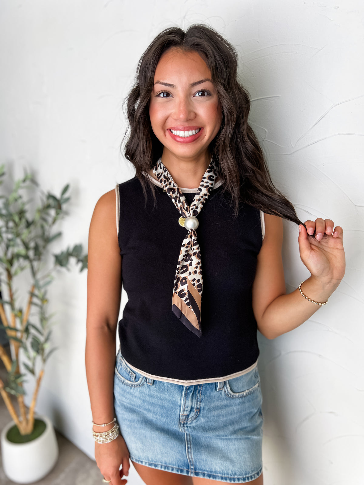 A portrait photograph of a smiling brunette woman holding a strand of her hair while posing against a textured off-white wall next to a potted olive plant. She is wearing a sleeveless black knit tank top made from a 70% viscose and 30% polyester blend, featuring light beige contrast ribbing along the crew neck, armholes, and bottom hem, paired with a light-wash denim skirt, styled with a leopard print silk neck scarf secured with a large pearl slide, a delicate gold beaded bracelet, and stacked beaded brace