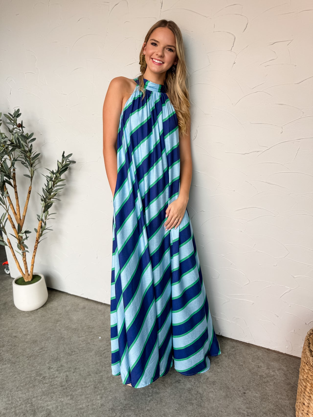 A full-length photograph of a smiling blonde woman standing against a textured white plaster wall, modeling a sweeping maxi dress made from an 80% rayon and 20% nylon blend. The sleeveless dress features a vibrant, multi-colored diagonal striped pattern in shades of dark navy, bright royal blue, light aqua blue, and kelly green. It is designed with a high, gathered halter neckline that falls into an ultra-flowy, voluminous tent silhouette reaching the floor. 
