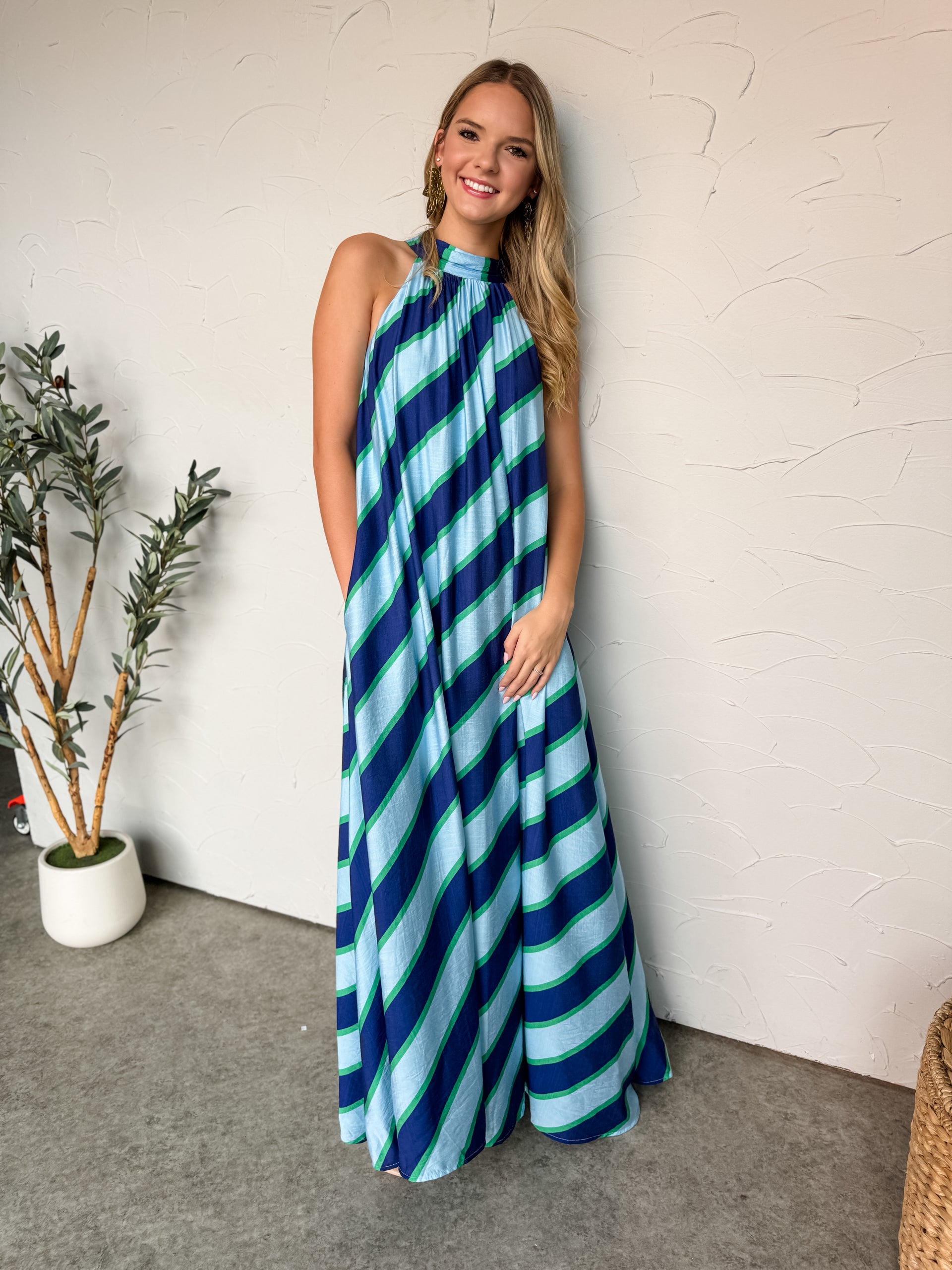 A full-length photograph of a smiling blonde woman standing against a textured white plaster wall, modeling a sweeping maxi dress made from an 80% rayon and 20% nylon blend. The sleeveless dress features a vibrant, multi-colored diagonal striped pattern in shades of dark navy, bright royal blue, light aqua blue, and kelly green. It is designed with a high, gathered halter neckline that falls into an ultra-flowy, voluminous tent silhouette reaching the floor. 