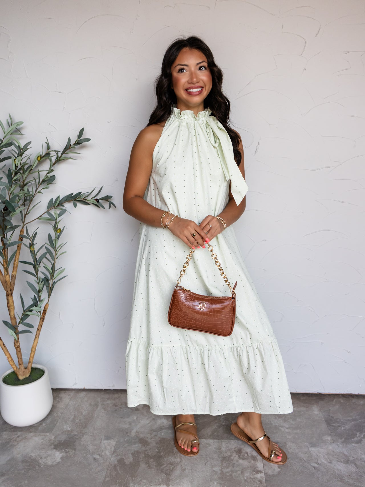A full-length photograph of a smiling woman with long wavy dark brown hair posing against a textured white wall while holding a brown croc-embossed handbag. She is wearing a sleeveless mint green midi dress with a green polka dot pattern made from 100% cotton fabric, featuring a high ruffled tie neckline and a tiered ruffle hem, styled with metallic flat sandals and gold bracelets.