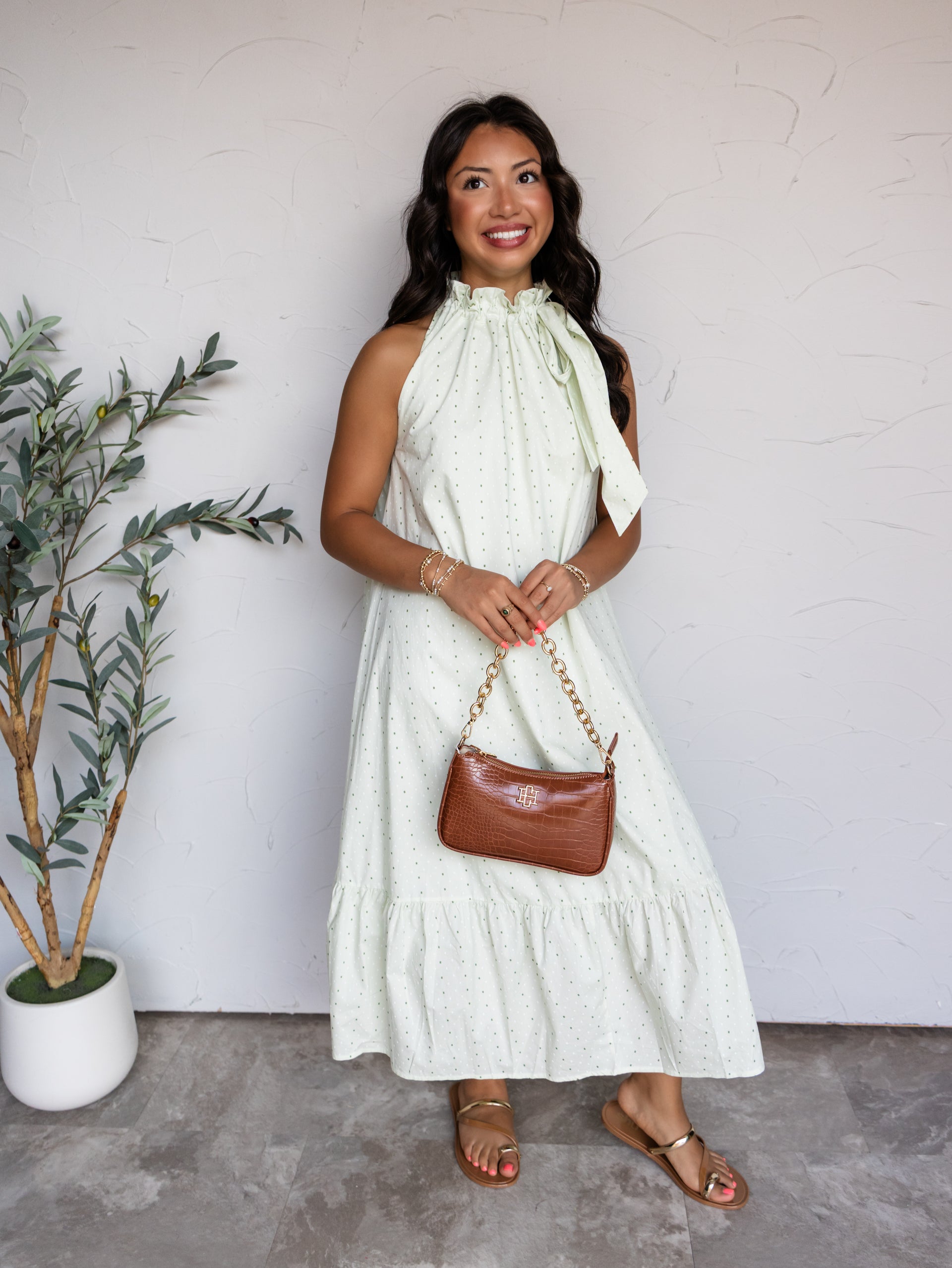 A full-length photograph of a smiling woman with long wavy dark brown hair posing against a textured white wall while holding a brown croc-embossed handbag. She is wearing a sleeveless mint green midi dress with a green polka dot pattern made from 100% cotton fabric, featuring a high ruffled tie neckline and a tiered ruffle hem, styled with metallic flat sandals and gold bracelets.