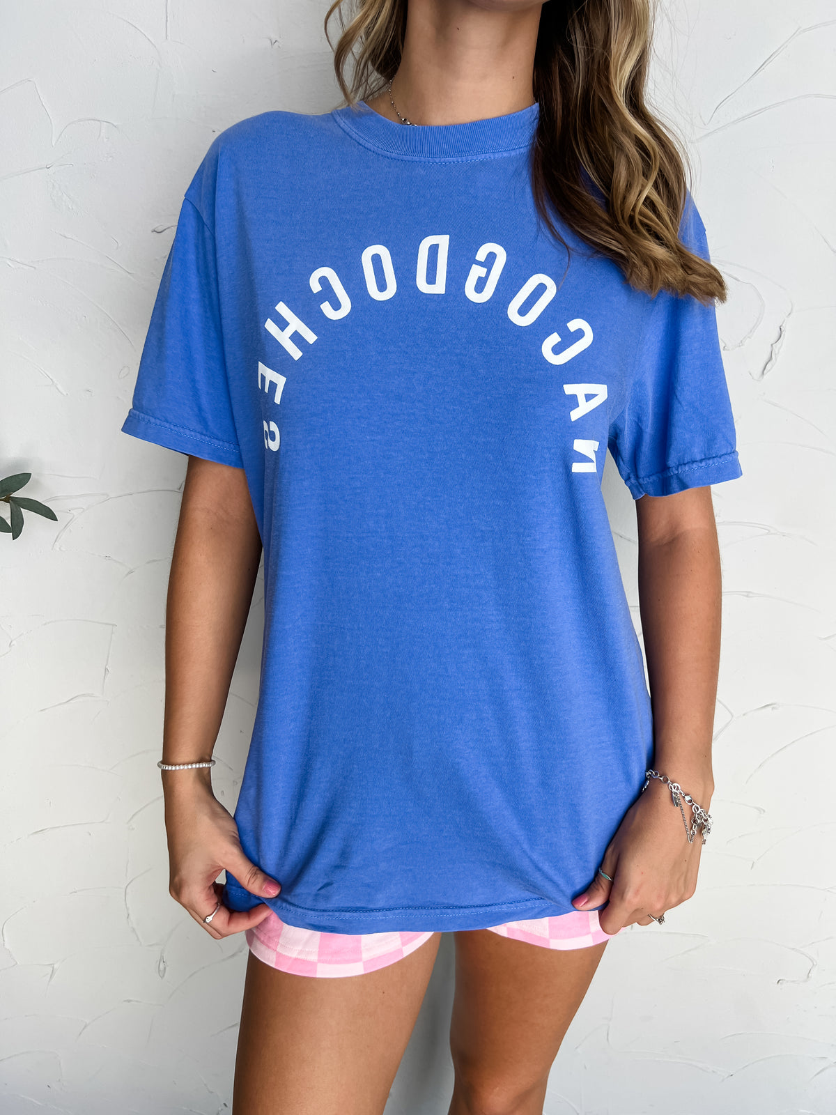A woman wearing a 100% cotton  heather blue graphic t-shirt with 'CAN COOCHES' printed in white arched text across the chest, paired with pink and white checkered shorts, standing against a white textured wall.