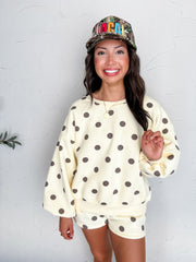 A portrait photograph of a smiling brunette woman posing against a textured white wall while holding a lock of her hair. She is wearing a cream-colored long-sleeved crewneck sweatshirt made from a 66% polyester and 45% cotton blend, decorated with dark brown polka dots, drop shoulders, and ribbed trim, paired with matching polka dot lounge shorts, styled with a camouflage trucker hat embroidered with "LOCAL" in colorful block lettering, a gold round pendant necklace, and a gold beaded bracelet.