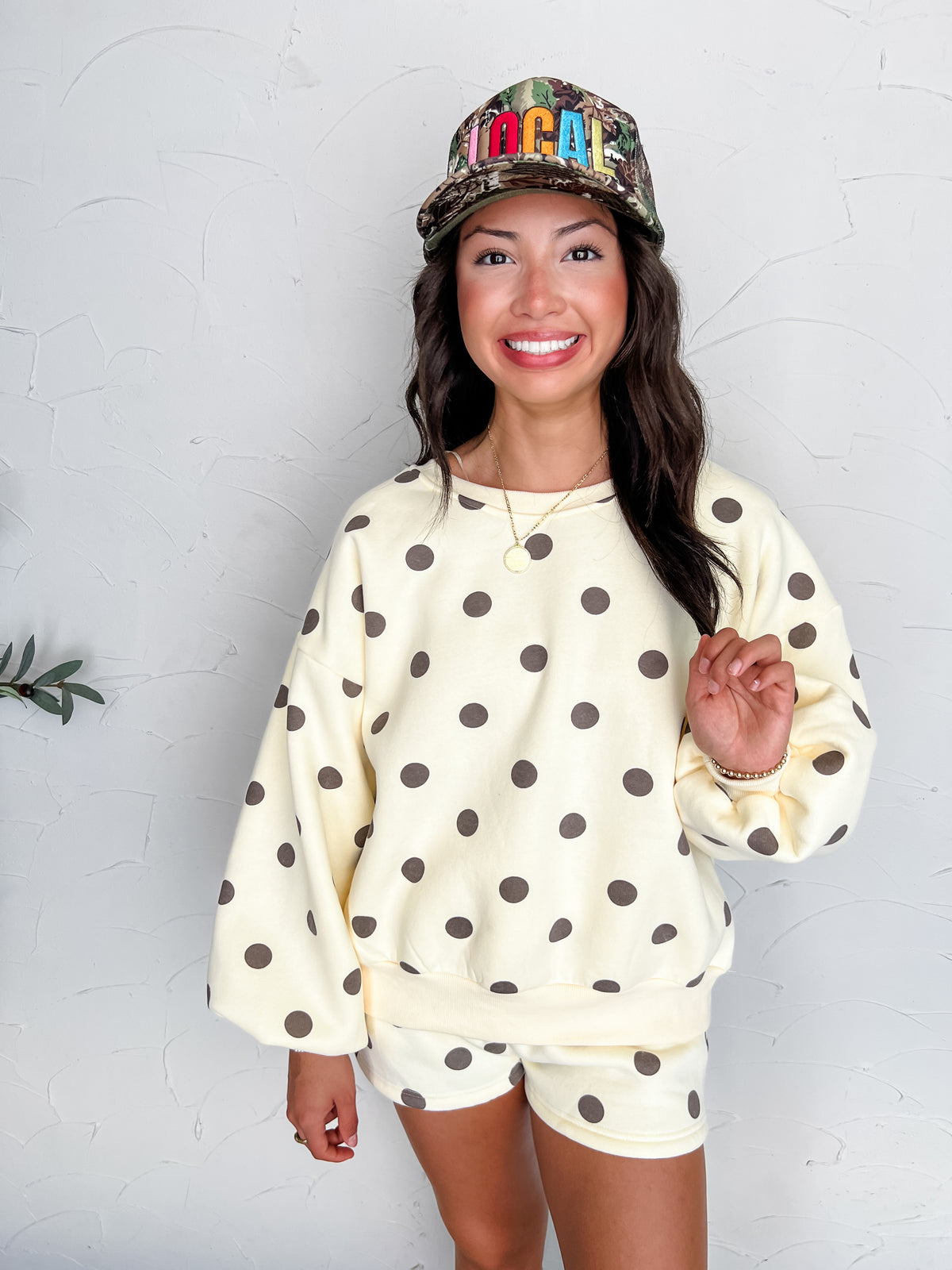 A portrait photograph of a smiling brunette woman posing against a textured white wall while holding a lock of her hair. She is wearing a cream-colored long-sleeved crewneck sweatshirt made from a 66% polyester and 45% cotton blend, decorated with dark brown polka dots, drop shoulders, and ribbed trim, paired with matching polka dot lounge shorts, styled with a camouflage trucker hat embroidered with "LOCAL" in colorful block lettering, a gold round pendant necklace, and a gold beaded bracelet.