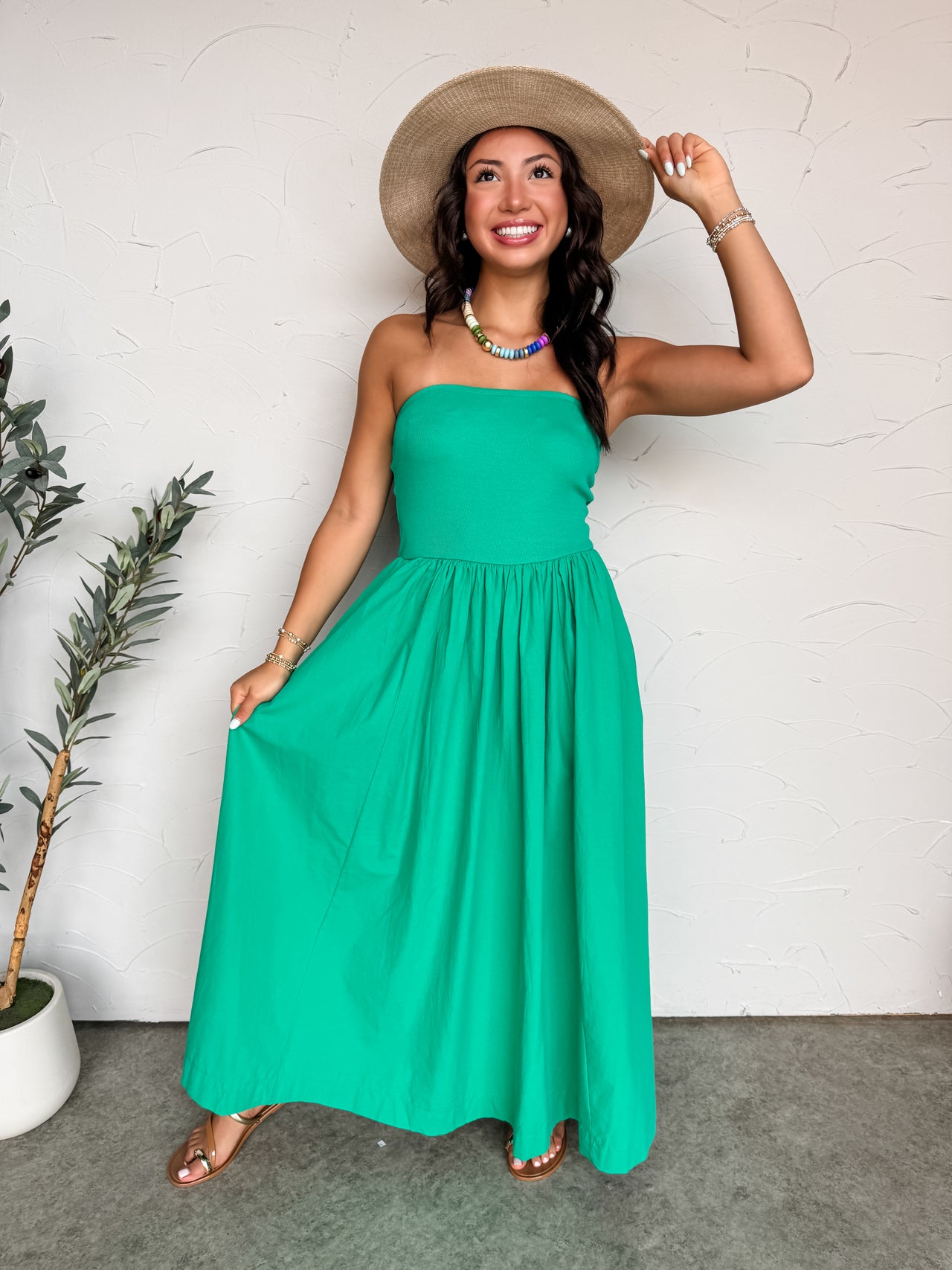 A full-length photograph of a smiling woman standing against a textured white wall, modeling a vibrant kelly green maxi dress made from 100% cotton. The dress features a strapless, tube-style bodice with a fine ribbed texture that meets a gathered, flowy maxi skirt at the waistline. She wears a multi-colored beaded collar necklace, stacked bracelets, and brown sandals.