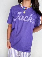 A person wearing a 100% Cotton heather purple short-sleeve t-shirt with 'Jacks' printed across the chest in a glittery white script, paired with lavender and white striped pants. The photo is cropped from the neck down, showing accessories like a colorful beaded necklace and bracelets against a white textured wall.