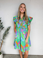 A full-length photograph of a smiling blonde woman standing against a textured white plaster wall, modeling a colorful mini dress made from 100% polyester. The dress features an eclectic, retro abstract print with bright shades of green, purple, orange, and hot pink. It is designed with double-layered flutter sleeves, a split V-neckline, and a high empire waist accented with solid green trim that flows into a tiered, loose skirt. 