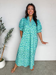 A full-length photograph of a smiling brunette woman with long wavy hair standing against a textured white plaster wall, modeling a loose, comfortable midi dress made from 100% cotton. The dress features a bright green and blue abstract leopard or animal print pattern throughout. It has short, voluminous puff sleeves and a split V-neckline, both accented with a contrasting green and white gingham checked trim.
