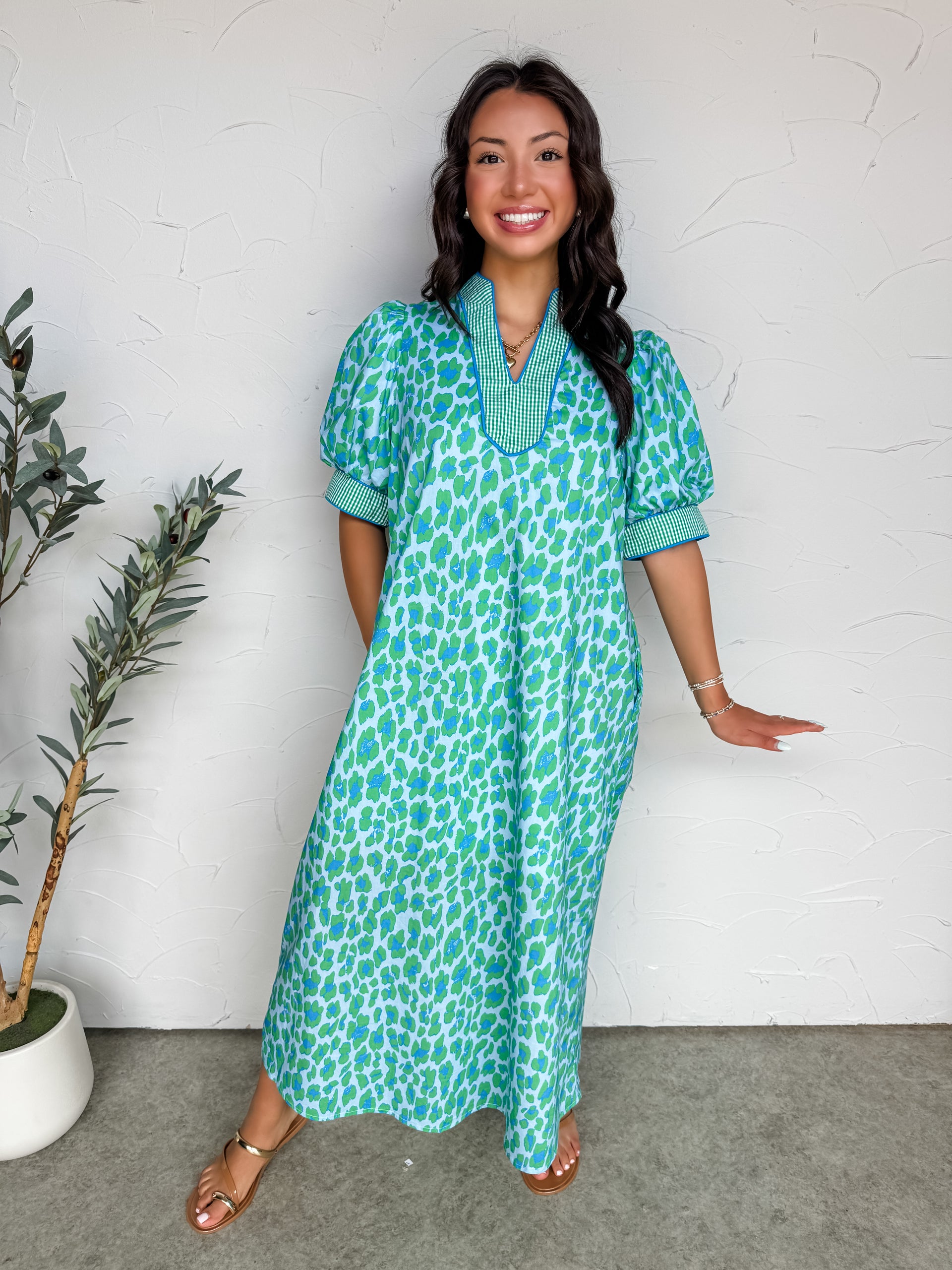 A full-length photograph of a smiling brunette woman with long wavy hair standing against a textured white plaster wall, modeling a loose, comfortable midi dress made from 100% cotton. The dress features a bright green and blue abstract leopard or animal print pattern throughout. It has short, voluminous puff sleeves and a split V-neckline, both accented with a contrasting green and white gingham checked trim.