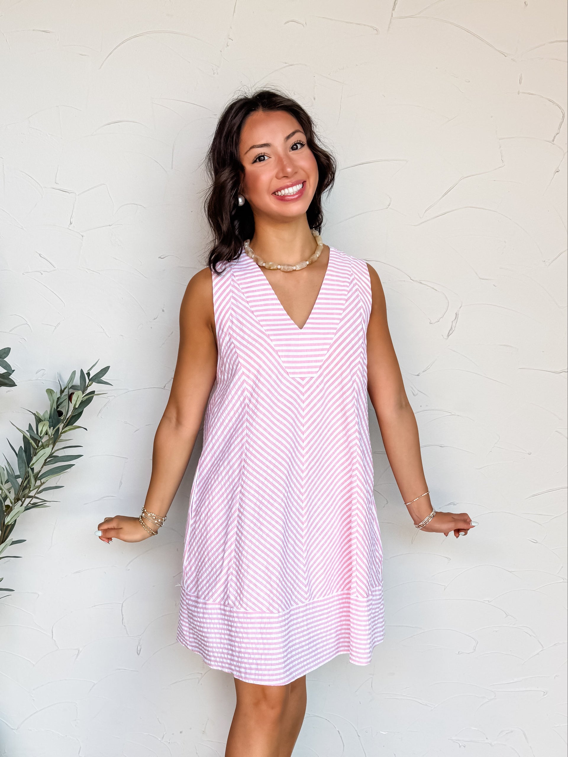 A full-length photograph of a smiling brunette woman posing against a textured white wall. She is wearing a sleeveless shift dress made from a 75% cotton and 25% polyester blend fabric featuring a pink and white multi-directional stripe pattern, a V-neckline, and a relaxed silhouette.
