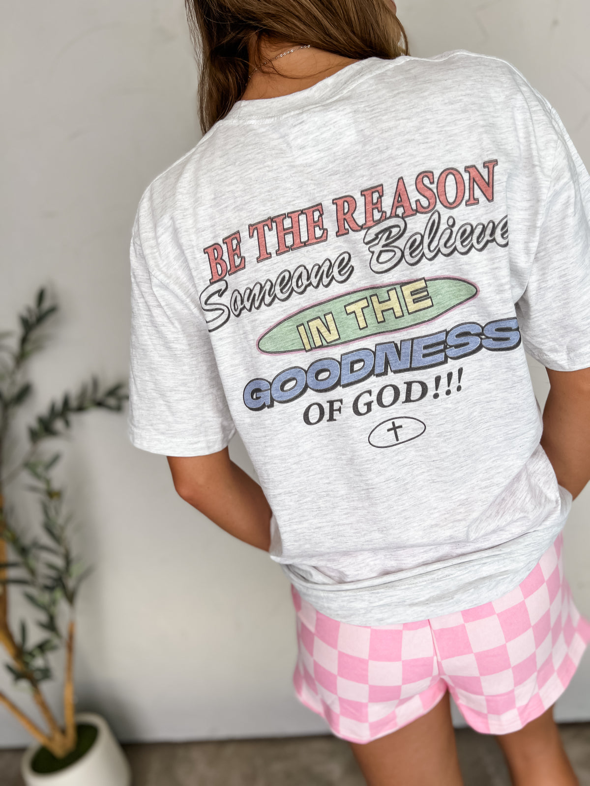 A person shown from behind wearing a 100% Cotton light heather gray graphic T-shirt with a large multicolor religious slogan on the back, paired with pink and white checkered shorts in an indoor setting.