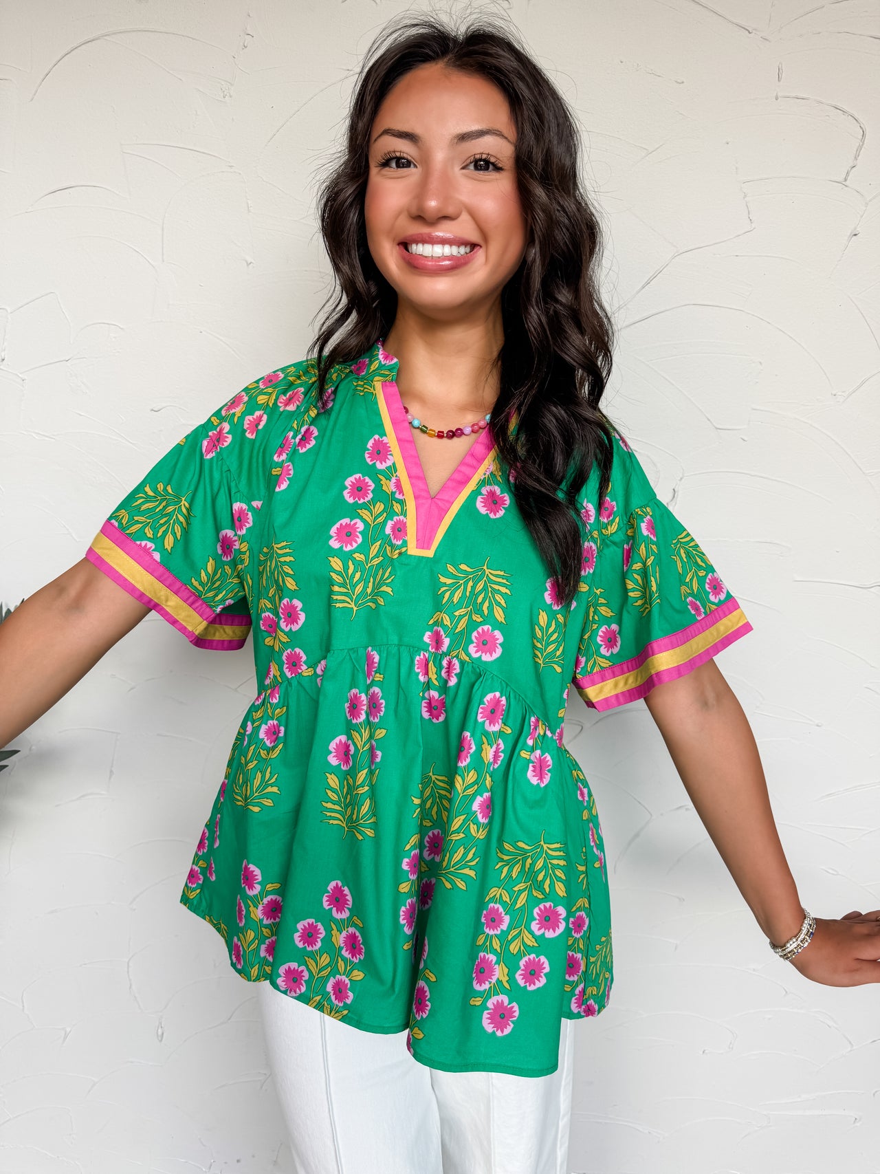 A full mid-shot photograph of a smiling brunette woman with wavy hair posing against a textured white plaster wall. She is wearing a bright green babydoll tunic blouse made from 100% cotton, which features an all-over block-print inspired pattern of pink flowers and golden-yellow stems. The top is designed with a split v-neckline and wide, relaxed short sleeves, both detailed with a contrasting pink and golden-yellow ribbon border. 