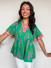 A full mid-shot photograph of a smiling brunette woman with wavy hair posing against a textured white plaster wall. She is wearing a bright green babydoll tunic blouse made from 100% cotton, which features an all-over block-print inspired pattern of pink flowers and golden-yellow stems. The top is designed with a split v-neckline and wide, relaxed short sleeves, both detailed with a contrasting pink and golden-yellow ribbon border. 