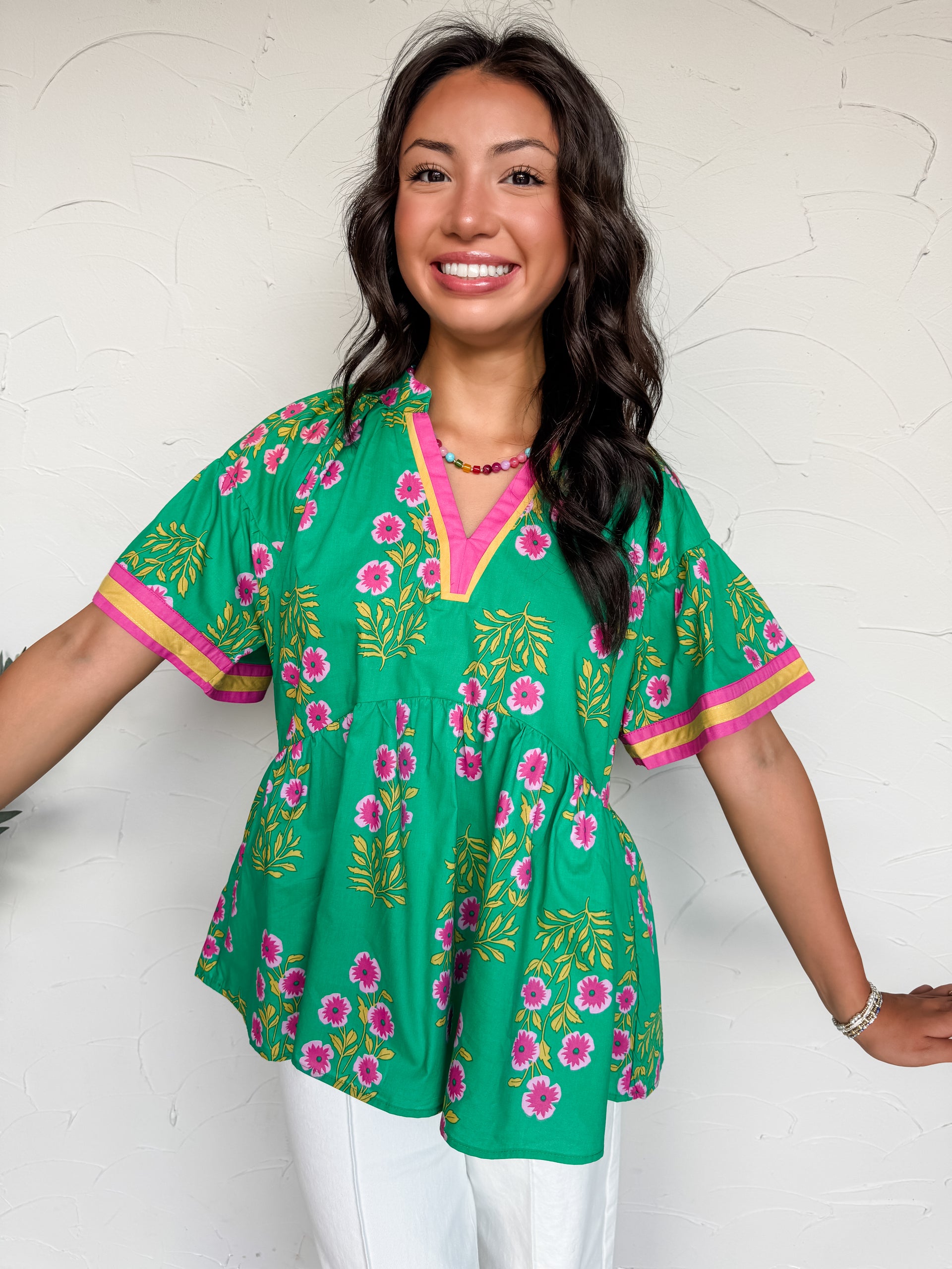 A full mid-shot photograph of a smiling brunette woman with wavy hair posing against a textured white plaster wall. She is wearing a bright green babydoll tunic blouse made from 100% cotton, which features an all-over block-print inspired pattern of pink flowers and golden-yellow stems. The top is designed with a split v-neckline and wide, relaxed short sleeves, both detailed with a contrasting pink and golden-yellow ribbon border. 