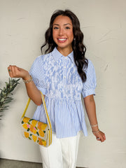 A close-up photograph focusing on the upper body of a smiling woman with dark wavy hair, standing against a white wall and modeling a stylish blouse made from a 75% polyester and 25% cotton blend. The top features a light blue and white vertical pinstripe pattern. It is detailed with short puff sleeves, a classic pointed collar, a partial front button placket, and a heavily smocked bodice that gives way to a flared peplum waist. 