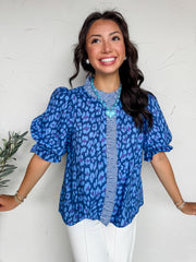 A close-up photograph focusing on the torso of a woman standing against a textured white plaster wall, modeling a vibrant blue blouse made from 100% cotton. The top features a bold, abstract leopard or animal print pattern in shades of medium blue, royal blue, and dark purple. It is designed with voluminous elbow-length puff sleeves that end in ruffled cuffs. The high neck and central button placket are prominently accented with a contrasting blue and white gingham checked ruffle trim. 