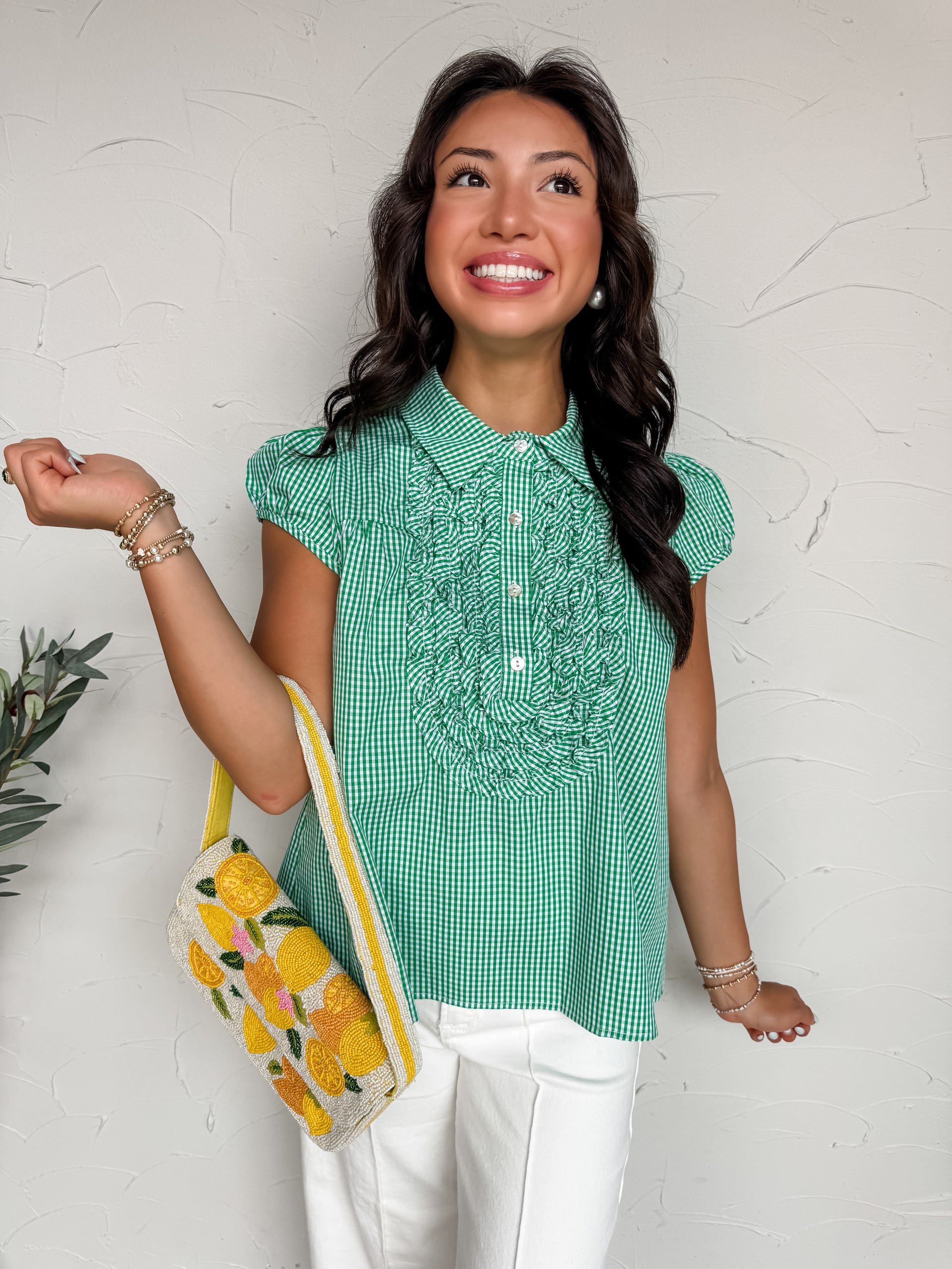 A close-up shot focusing from the neck down of a woman wearing a green and white gingham tunic top made of 100% cotton. The blouse features a classic pointed collar, a button-front closure, cap sleeves, and a prominent decorative bib composed of layered ruffles on the chest. She is paired with white pants and carries a heavily beaded handbag adorned with a vibrant yellow lemon pattern.