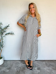 A full-length photograph of a smiling blonde woman with long wavy hair posing with one hand on her hip and one hand in her pocket against a textured off-white wall next to a potted olive plant. She is wearing a short-sleeved midi-length shirt dress made from a 68% polyester, 23% cotton, and 9% rayon blend in a black and white gingham pattern, featuring a pointed open collar, a partial button placket, side pockets, and side hem slits, styled with black leather slide sandals with gold buckle hardware, a simpl