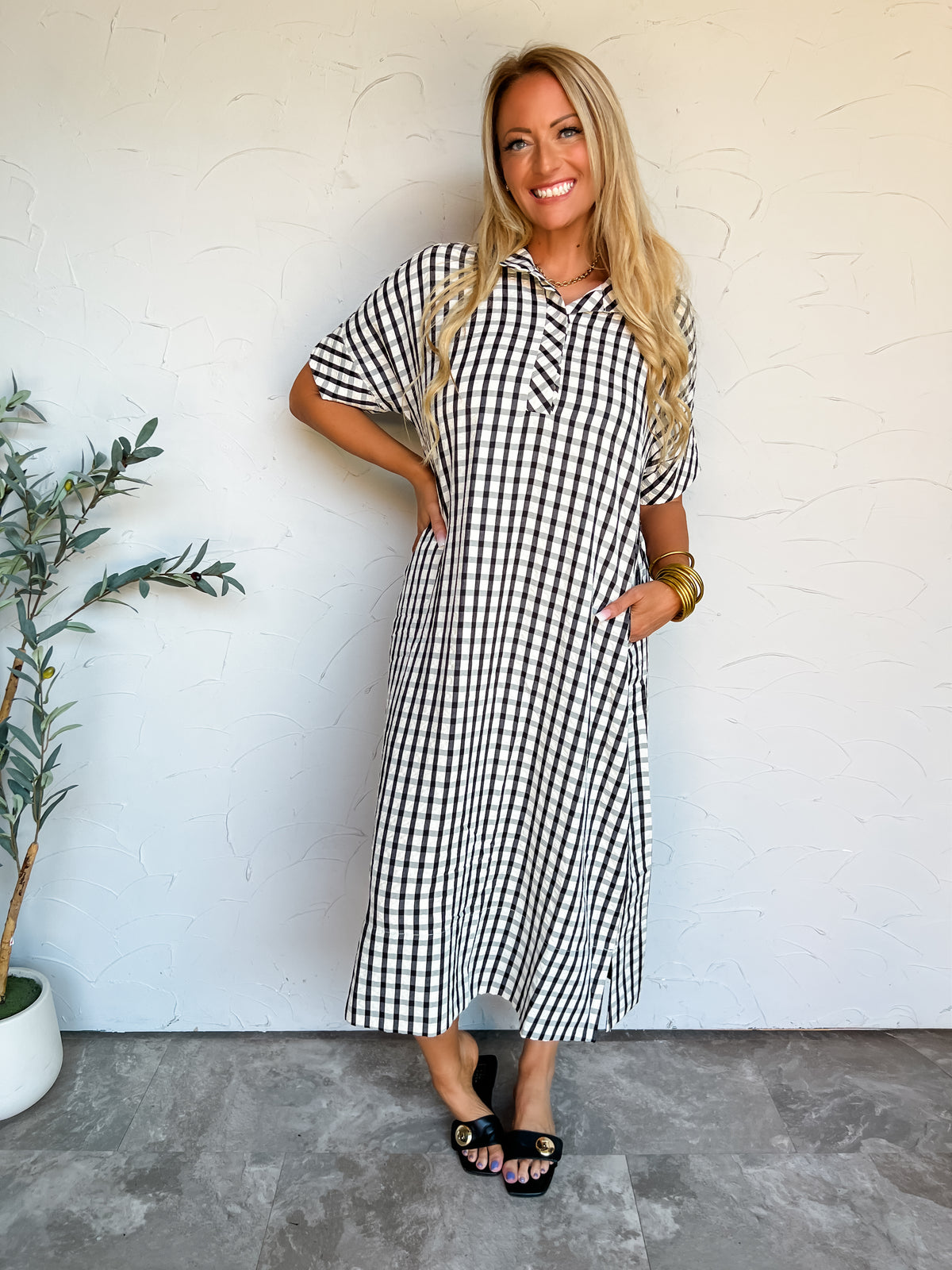 A full-length photograph of a smiling blonde woman with long wavy hair posing with one hand on her hip and one hand in her pocket against a textured off-white wall next to a potted olive plant. She is wearing a short-sleeved midi-length shirt dress made from a 68% polyester, 23% cotton, and 9% rayon blend in a black and white gingham pattern, featuring a pointed open collar, a partial button placket, side pockets, and side hem slits, styled with black leather slide sandals with gold buckle hardware, a simpl