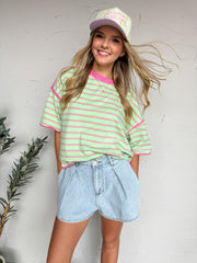 A mid-shot photograph of a smiling blonde woman with her hair blowing in the wind, posing against a textured white plaster wall. She is wearing an oversized, casual crewneck t-shirt made from a 95% cotton and 5% spandex blend. The knit top features a horizontal stripe pattern in lime green and hot pink, with a contrasting hot pink ribbed neckline and exposed reverse stitching at the dropped shoulders. 