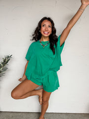 A full-length photograph of a smiling brunette woman posing on one leg against a textured white wall. She is wearing a matching two-piece lounge set in a vibrant green mineral-wash finish, featuring an oversized t-shirt with a chest pocket and matching high-waisted shorts.