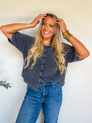 A portrait photograph of a smiling blonde woman posing with both hands on her hair against a textured off-white wall. She is wearing a dark charcoal blue ribbed knit short-sleeve cardigan made from 100% cotton, featuring a button-up front and a crew neckline, tucked into medium-wash high-waisted jeans, styled with stacked gold bangles, a gold chain necklace, and rings.