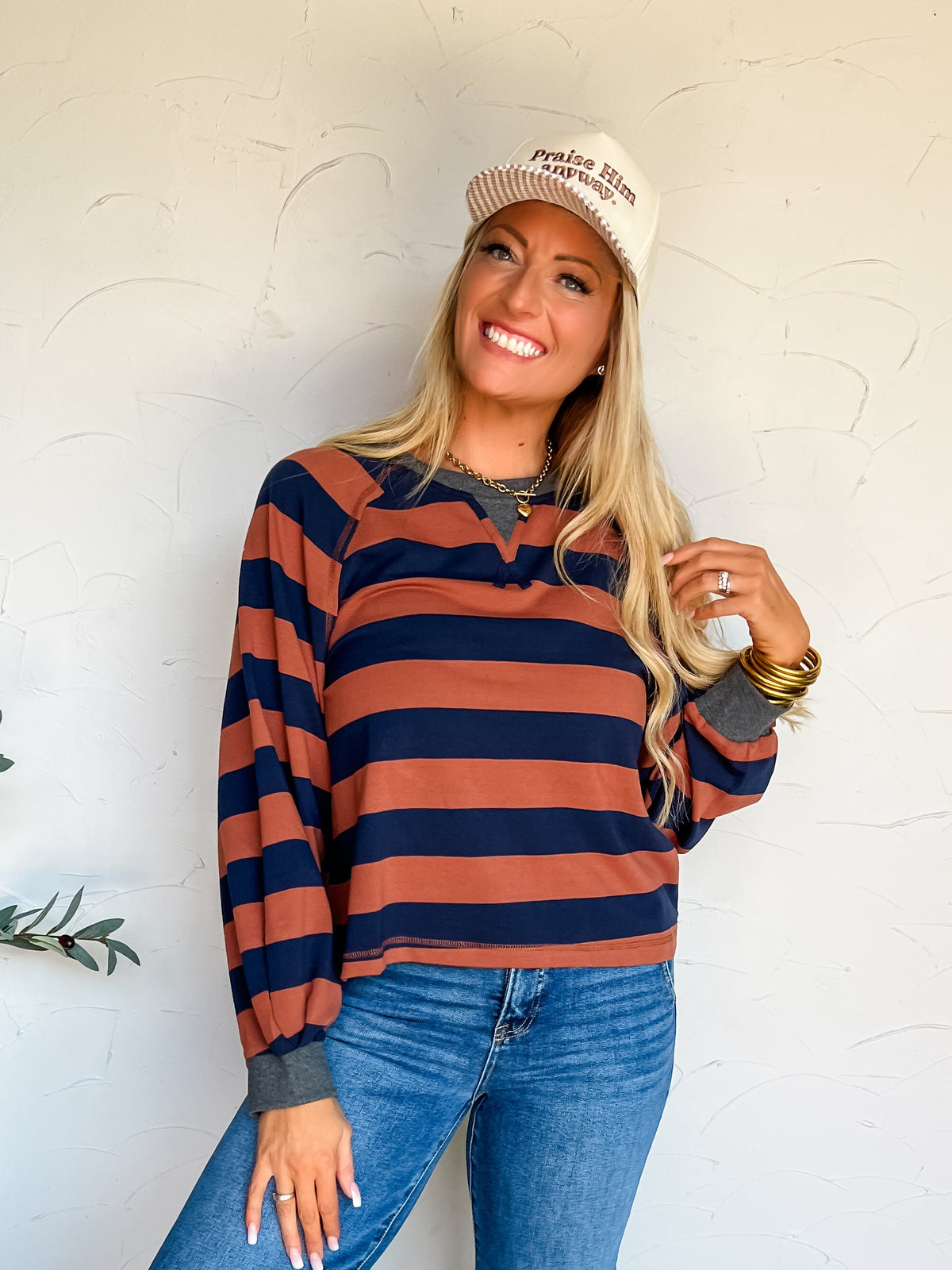 She is wearing a long-sleeved top made from a 93% cotton and 7% spandex blend in a rust brown and navy blue horizontal stripe print, featuring a charcoal grey collar notch and wrist cuffs, paired with medium-wash cuffed wide-leg denim jeans, tan leather tassel slide loafers, a cream baseball cap embroidered with "Praise Him anyway.", stacked gold bangles, and a gold chain necklace.