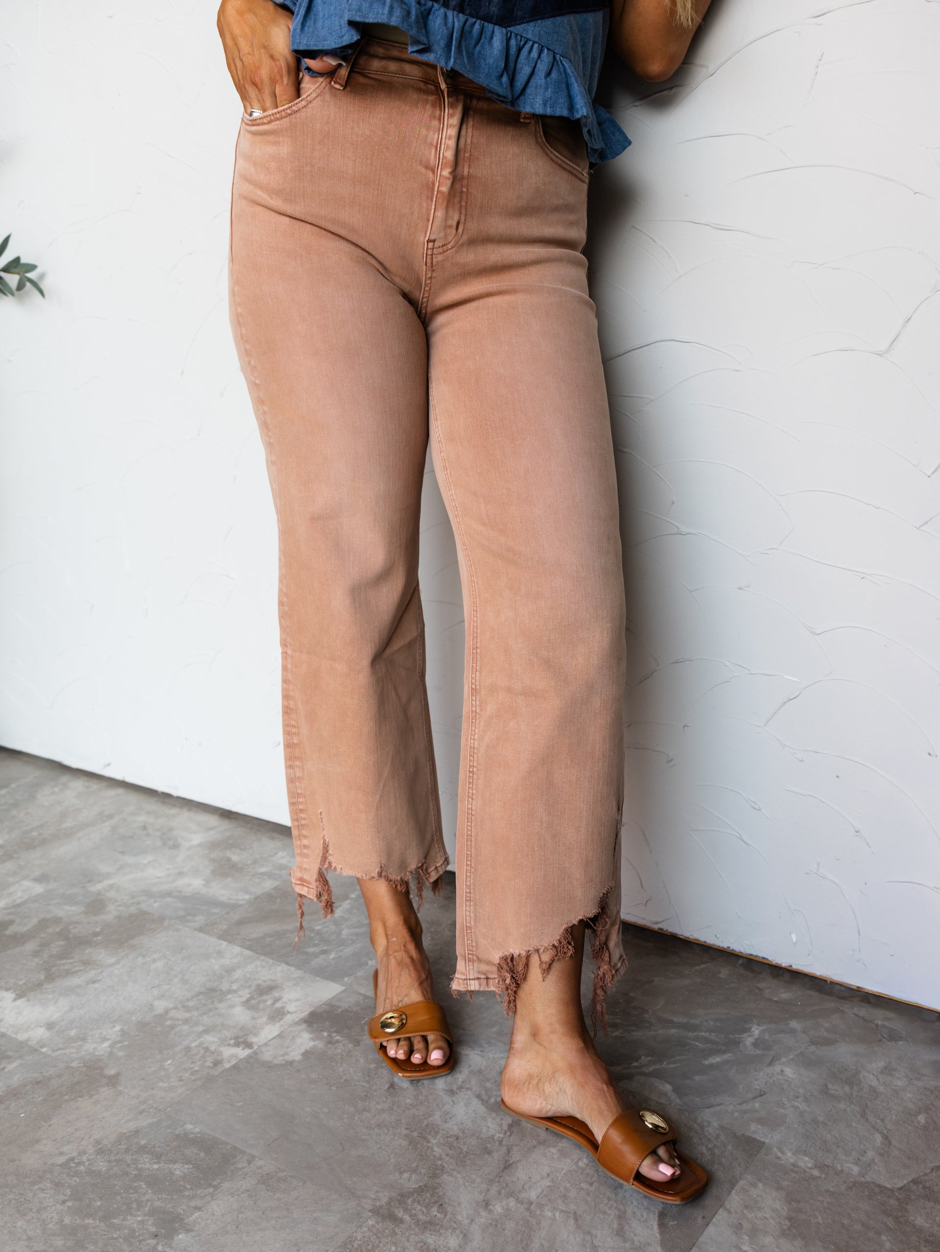 A full-length photograph focused from the waist down of a woman posing with one hand in her pocket against a textured white wall. She is wearing blush tan wide-leg jeans made from a blend of 64% cotton, 28% lyocell, 6% polyester, and 2% spandex, featuring a heavily frayed distressed raw hem, paired with a blue denim peplum top and cognac brown leather slide sandals.