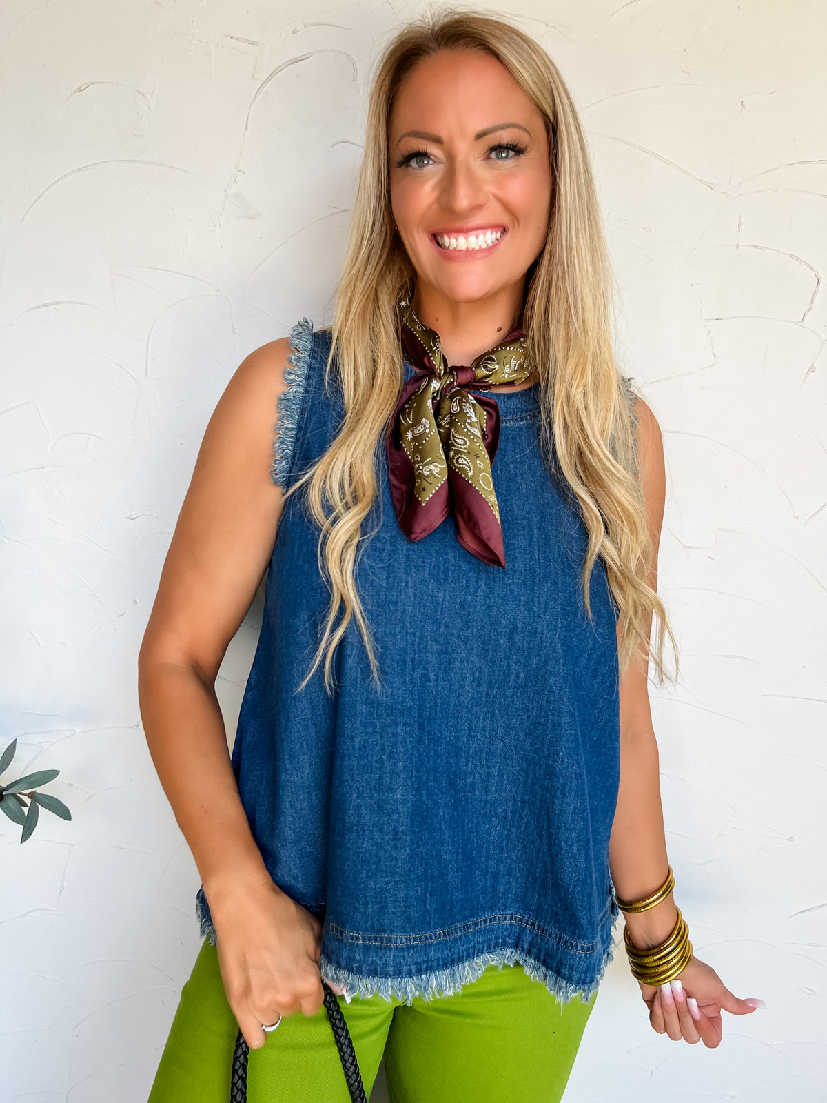 A portrait photograph of a smiling blonde woman with long wavy hair posing against a textured off-white wall. She is wearing a sleeveless medium-wash denim tank top made from a 75% cotton and 25% polyester blend, featuring raw frayed trim along the armholes and hemline, paired with lime green pants, styled with an olive green and burgundy paisley neck scarf, stacked gold bangles, and holding a black woven bag strap.