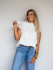 A three-quarter length photograph of a smiling woman with long blonde hair holding the ruffle on her sleeve while posing against a textured white wall. She is wearing a white floral lace sleeveless top made from 100% polyester fabric, featuring a high ruffled mock neckline and flutter lace sleeves, tucked into medium-wash denim jeans and styled with stacked gold bangles