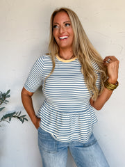 A portrait photograph of a smiling blonde woman with long wavy hair posing against a textured off-white wall next to a potted olive plant. She is wearing a slate blue and white horizontally striped peplum top made from a 92% polyester and 8% spandex blend, featuring a soft yellow crew neckband, elbow-length sleeves, and a flared ruffled hem, paired with light-wash denim jeans with front patch pockets, styled with stacked gold bangles, a gold chain necklace, and a diamond ring.