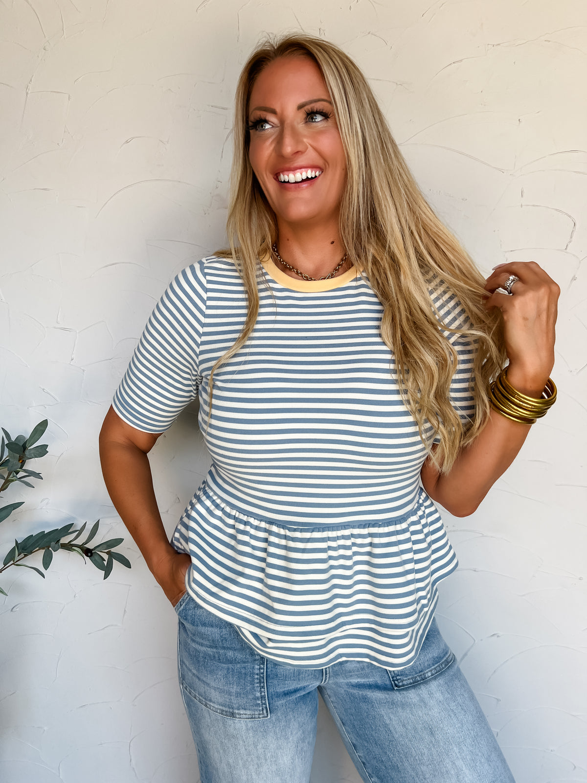 A portrait photograph of a smiling blonde woman with long wavy hair posing against a textured off-white wall next to a potted olive plant. She is wearing a slate blue and white horizontally striped peplum top made from a 92% polyester and 8% spandex blend, featuring a soft yellow crew neckband, elbow-length sleeves, and a flared ruffled hem, paired with light-wash denim jeans with front patch pockets, styled with stacked gold bangles, a gold chain necklace, and a diamond ring.