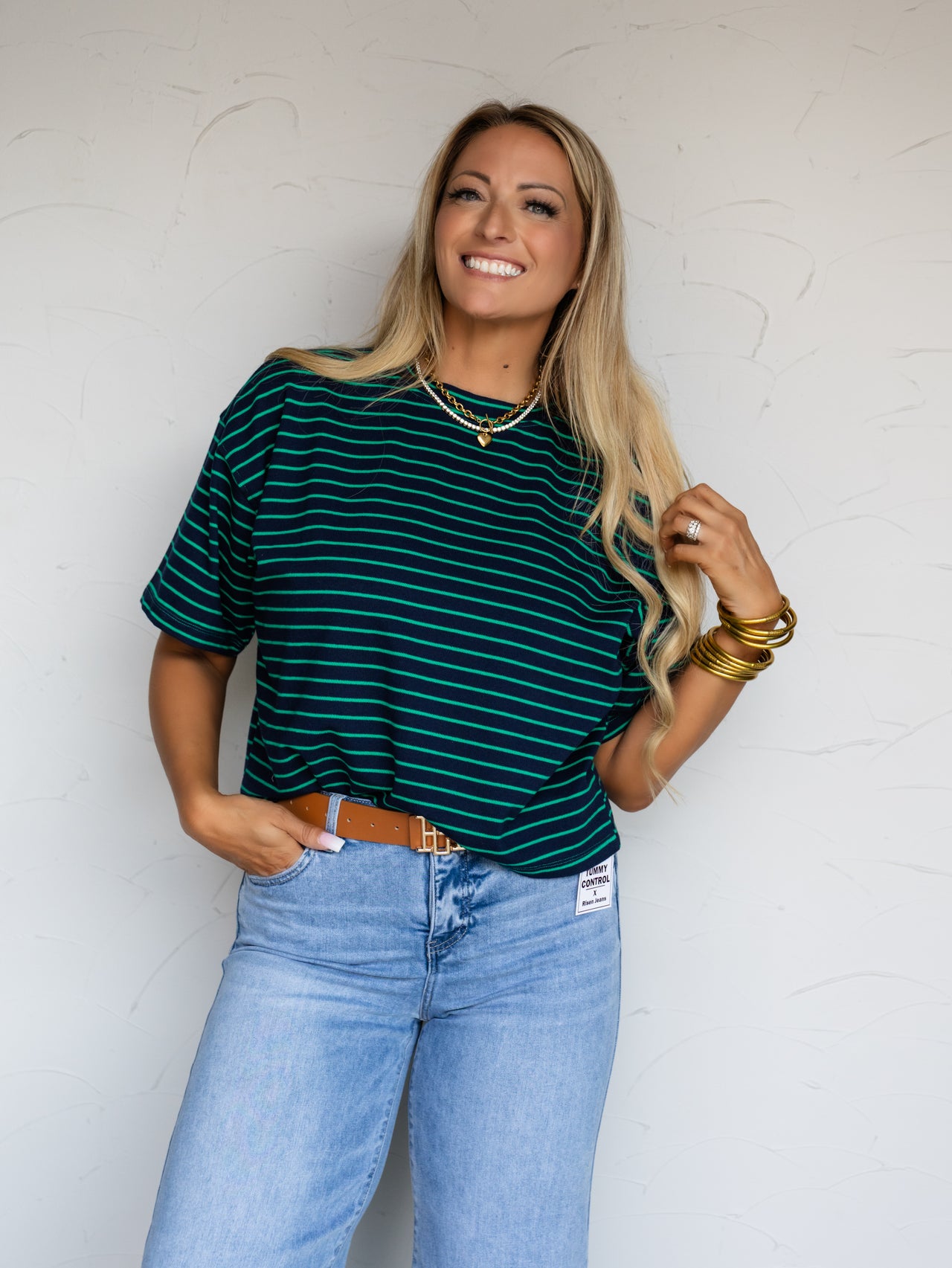 A three-quarter length photograph of a smiling woman with long blonde hair holding a strand of her hair while posing with one hand in her pocket against a textured white wall. She is wearing a navy blue and kelly green horizontal striped short-sleeved t-shirt made from 100% cotton fabric featuring cuffed sleeves and a crew neck, tucked into light-wash denim jeans with a brown belt and styled with gold chain necklaces and stacked gold bangles.