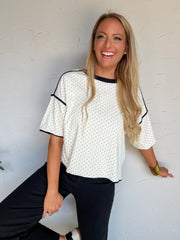 A three-quarter length photograph of a smiling blonde woman leaning against a textured white wall while lifting one leg. She is wearing a white oversized short-sleeve top made from a 95% cotton and 5% spandex blend, featuring a black micro-polka dot pattern, a black crew neckband, and black piping along the drop-shoulder sleeves and seams, paired with black pants, metallic silver shoes, thick gold bangle bracelets, and small stud earrings.
