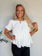 A portrait photograph of a smiling blonde woman with long wavy hair posing against a textured off-white wall next to a potted olive plant with one hand on her hip and touching her hair with the other. She is wearing a short-sleeved white top made from 100% cotton, featuring tone-on-tone embroidered floral geometric accents, puff sleeves with elastic cuffs, a round neckline, and a tiered peplum bodice, paired with black pants, styled with a pearl necklace with a gold heart pendant, stacked gold bangles, and 