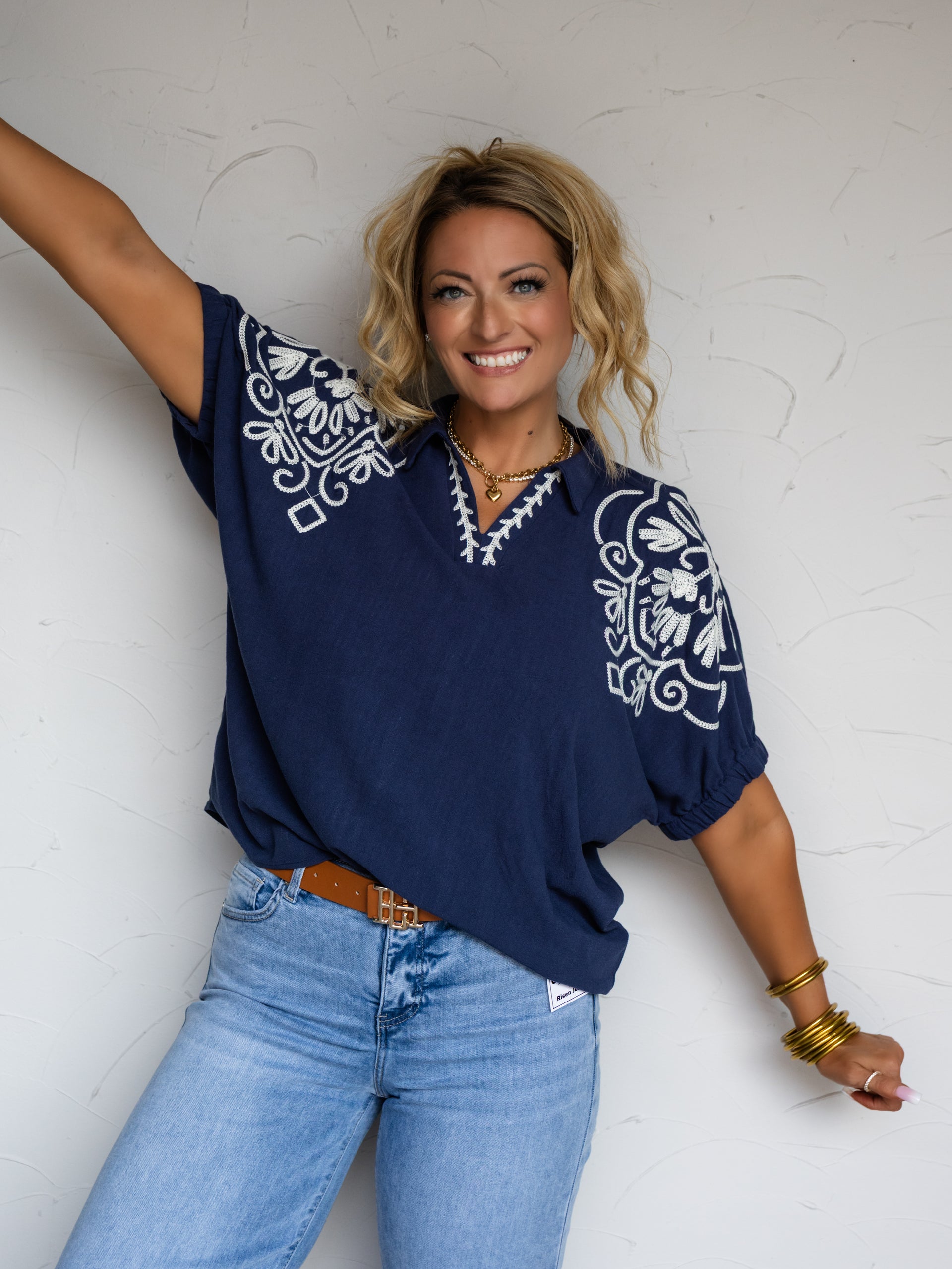 A waist-up photograph of a smiling woman with wavy blonde hair posing enthusiastically with arms outstretched against a textured white wall. She is wearing a dark navy blue short-sleeved blouse made from a 55% linen and 45% cotton blend, featuring intricate white floral embroidery across the shoulders and a split V-neckline, tucked into light-wash denim jeans with a cognac brown belt and styled with a gold chain necklace and stacked gold bangles.
