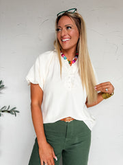 A three-quarter length photograph of a smiling blonde woman with long straight hair looking off to the side while touching her hair against a textured white wall. She is wearing a short-sleeved cream off-white top made from a 70% rayon and 30% nylon blend with a crinkle-textured weave, featuring a split V-neckline with a ruffle-trimmed half-button Henley placket and relaxed short sleeves, front-tucked into high-waisted dark olive green pants with a tan leather belt, styled with a colorful beaded necklace, g