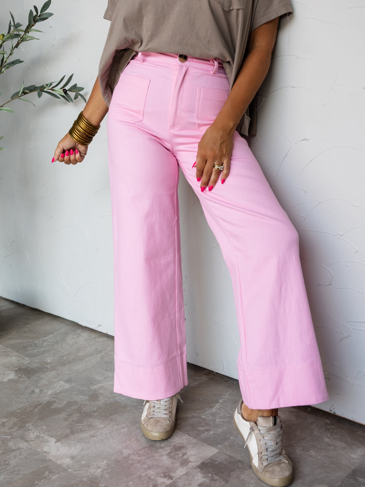 A full-length photograph focused from the waist down of a woman posing against a textured white wall. She is wearing bright pink high-waisted wide-leg pants made from a blend of 70% cotton, 28% polyester, and 2% spandex, featuring front patch pockets, paired with a tucked-in taupe t-shirt, low-top distressed sneakers, and gold bangles.