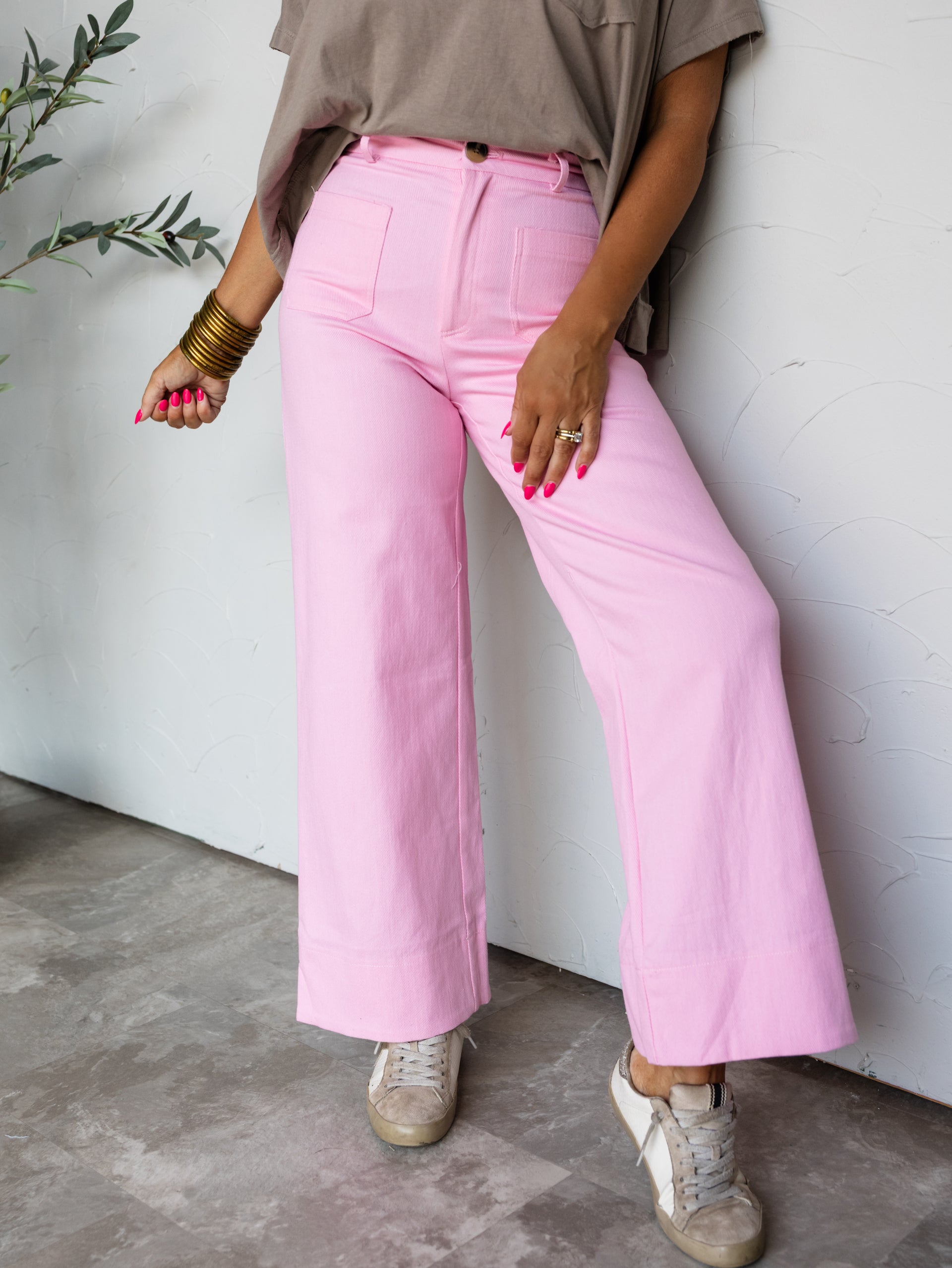 A full-length photograph focused from the waist down of a woman posing against a textured white wall. She is wearing bright pink high-waisted wide-leg pants made from a blend of 70% cotton, 28% polyester, and 2% spandex, featuring front patch pockets, paired with a tucked-in taupe t-shirt, low-top distressed sneakers, and gold bangles.
