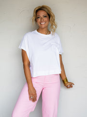 A three-quarter length photograph of a smiling woman with styled blonde hair in an updistressed pose against a textured white wall. She is wearing a crisp white short-sleeved t-shirt made from 100% cotton fabric featuring a chest pocket, cuffed sleeves, and a crew neck, paired with light pink pants and styled with gold chain necklaces and stacked gold bangles.