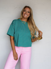 A three-quarter length photograph of a smiling woman with long blonde hair touching her hair while posing against a textured white wall. She is wearing a teal green and pink horizontal striped short-sleeved t-shirt made from 100% cotton fabric featuring cuffed sleeves and a crew neck, paired with light pink pants and styled with gold chain necklaces and stacked gold bangles.