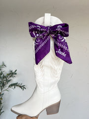 A white faux-leather cowboy boot with a brown block heel, featuring a large purple bandana bow with white western print tied around the collar.