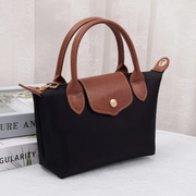 A black canvas tote bag with brown textured leather handles and trim, featuring a gold-tone snap button closure on the front flap. The bag is styled on top of stacked books against a soft, light-colored curtain background.