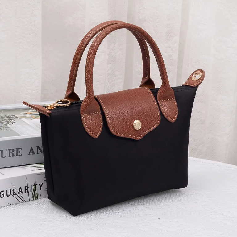 A black canvas tote bag with brown textured leather handles and trim, featuring a gold-tone snap button closure on the front flap. The bag is styled on top of stacked books against a soft, light-colored curtain background.
