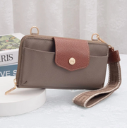 A taupe-brown crossbody wallet with a brown textured flap, gold-tone hardware, and a matching patterned wristlet strap, styled on a white pedestal with a book in the background.