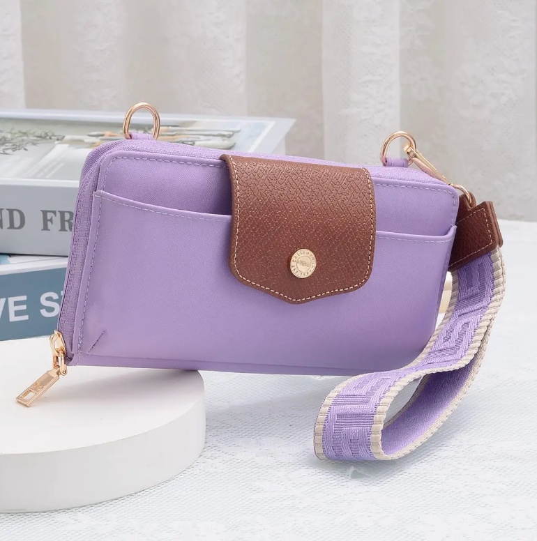 A lavender purple crossbody phone wallet bag with a brown textured flap and a matching purple patterned strap, displayed on a white round pedestal with books in the background.