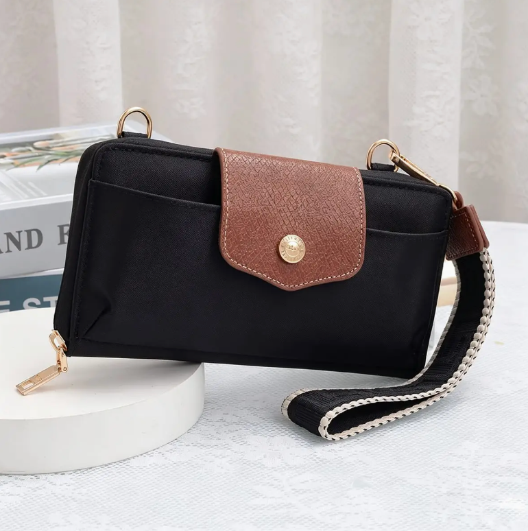A black nylon crossbody wallet with a brown textured flap and gold-tone hardware, displayed on a white pedestal with a patterned curtain in the background.