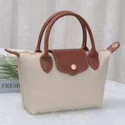 A small structured handbag with a cream-colored body and brown handles, flap, and trim, shown on a white textured surface with a soft curtain background.