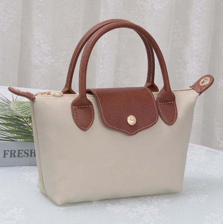 A small structured handbag with a cream-colored body and brown handles, flap, and trim, shown on a white textured surface with a soft curtain background.