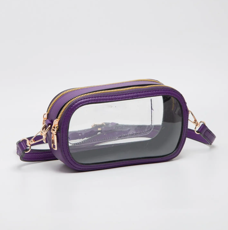 A purple faux-leather crossbody bag with a clear plastic front window and gold-tone hardware, shown on a light gray studio background.